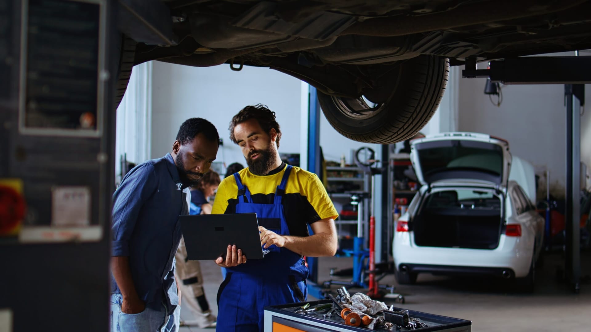 Trained mechanic and customer standing underneath car in garage using laptop to order new parts after finding defects. Employee assisting african american client by looking for components online